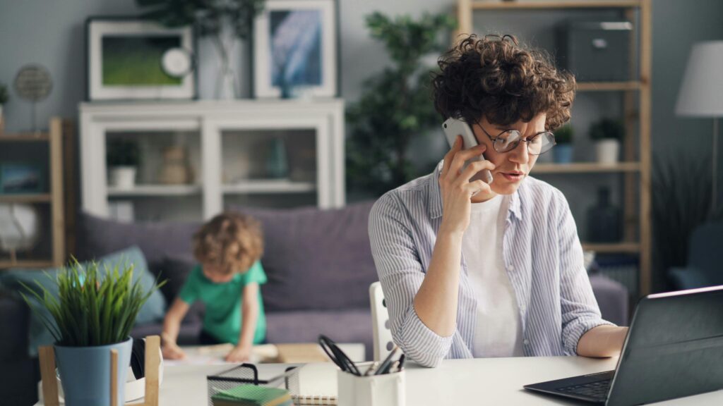 Parent working from home on a laptop and phone while her child plays nearby